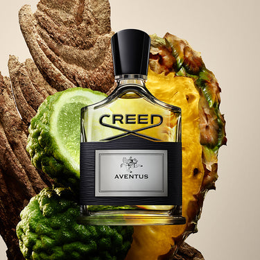 Creed Aventus perfume bottle surrounded by pineapple and lime slices on a beige background